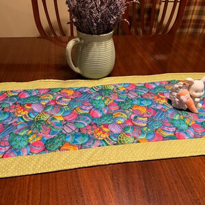 Puede incluir: Un camino de mesa verde con un borde amarillo y un patr&oacute;n de huevos de Pascua coloridos. Una figurita de conejo de cer&aacute;mica blanca est&aacute; sentada en el camino de mesa.