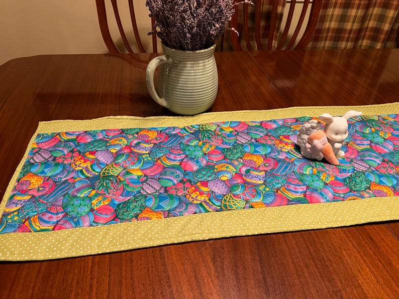 Puede incluir: Un camino de mesa verde con un borde amarillo y un patr&oacute;n de huevos de Pascua coloridos. Una figurita de conejo de cer&aacute;mica blanca est&aacute; sentada en el camino de mesa.