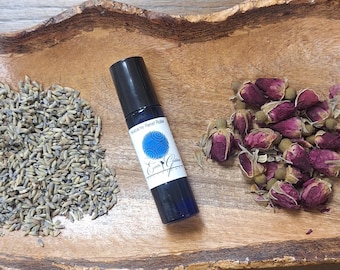 Puede incluir: Un cuenco de madera lleno de flores de lavanda secas, capullos de rosa y una pequeña botella de vidrio azul con un aplicador de bola rodante. La botella tiene una etiqueta blanca con letras azules que dice "Love & Grace".