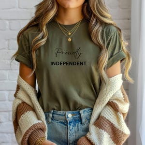 May include: An olive green t-shirt with the words "Proudly Independent" in black script. The shirt is worn with blue jeans and a cream and brown striped cardigan. Gold necklaces complete the casual look.