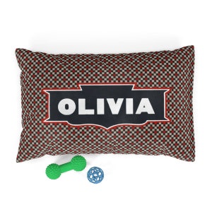 May include: A red and black checkered dog bed with the name "OLIVIA" in white text on a black banner. The bed is accompanied by a green rubber bone and a blue rubber ball.