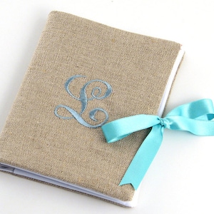May include: A burlap-covered journal with a white border and a light blue ribbon tied around it. The journal has a light blue embroidered letter "L" on the front.
