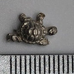 May include: A small, silver-colored turtle figurine, approximately 1 cm in length. The turtle is in a walking position with its legs outstretched.