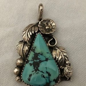 May include: A turquoise pendant with silver leaf and flower accents. The turquoise is a vibrant blue-green color with black veins.