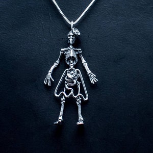 May include: A silver skeleton pendant necklace with a detailed anatomical design, including visible organs and bones. The skeleton is wearing a skirt and has a flower on its chest.