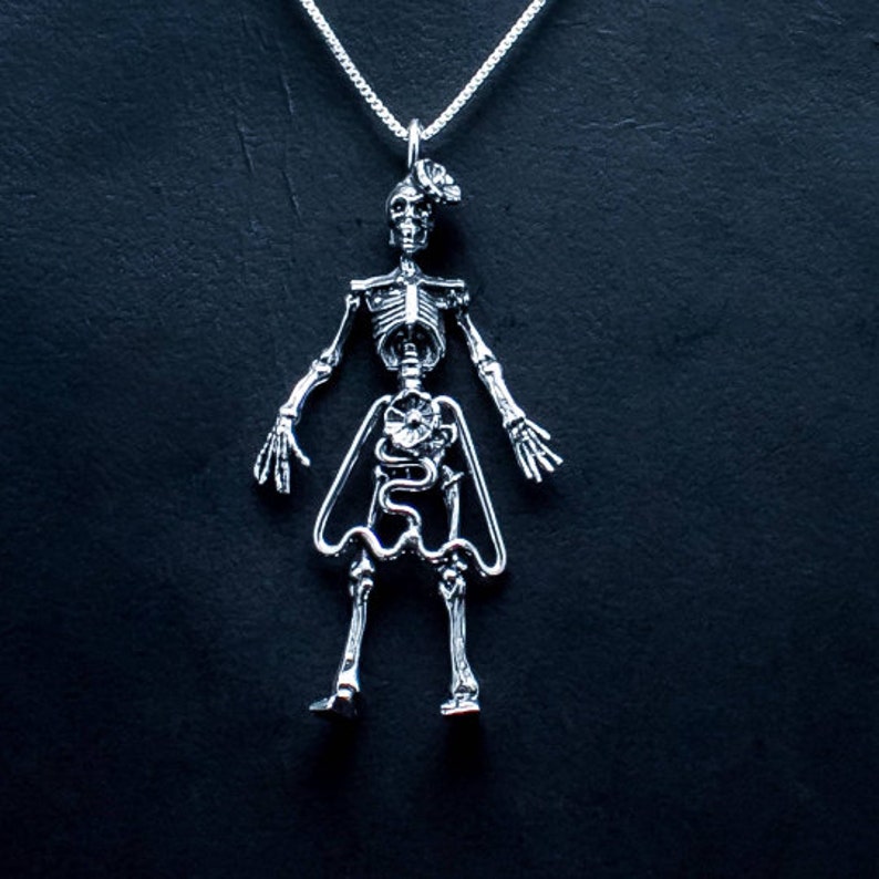May include: A silver skeleton pendant necklace with a detailed anatomical design, including visible organs and bones. The skeleton is wearing a skirt and has a flower on its chest.