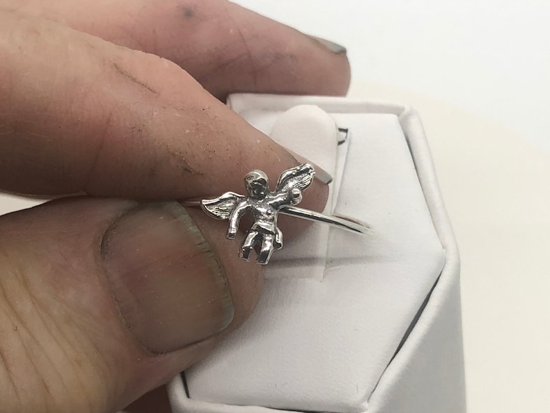 May include: A silver ring with a cherub design. The cherub is holding a heart in its hands.