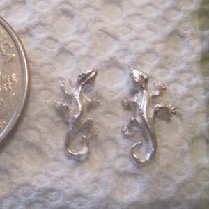 May include: Two silver-colored gecko lizard earrings. The earrings are about the size of a US quarter.