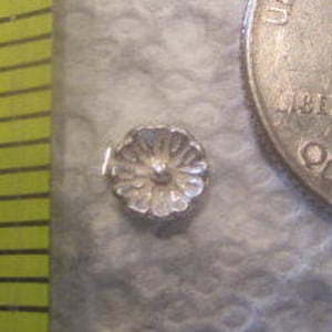 May include: A small, silver-colored flower-shaped bead. The bead is next to a U.S. quarter for size comparison. The bead's diameter appears to be less than 0.25 inches.