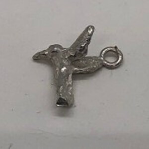 May include: A small, silver-colored hummingbird charm with outstretched wings. The charm has a detailed, textured surface and a small loop for attaching to a necklace or bracelet. The hummingbird is depicted in mid-flight.