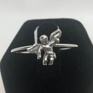 May include: A silver ring with a small angel figurine. The angel has wings and is holding a small object in its hand.