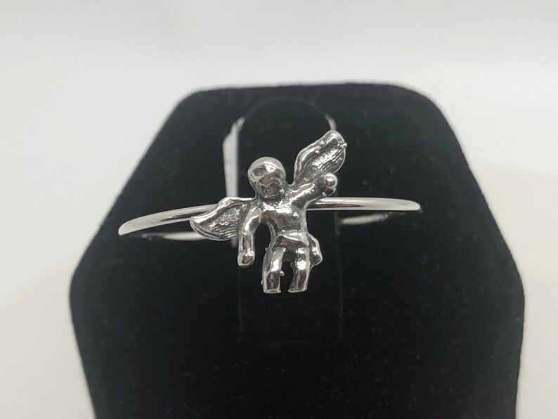 May include: A silver ring with a small angel figurine. The angel has wings and is holding a small object in its hand.