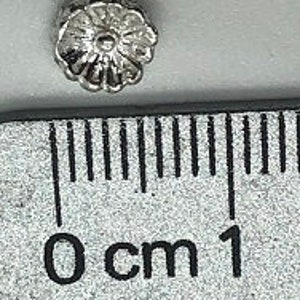 May include: A small, silver-colored flower-shaped button with a raised center. The button is about 0.5 inches in diameter.