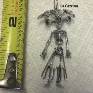 May include: Silver pendant featuring a skeletal figure with a large head and a wide brimmed hat. The pendant is labeled "La Catrina".