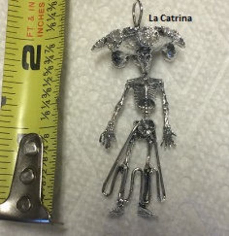 May include: Silver pendant featuring a skeletal figure with a large head and a wide brimmed hat. The pendant is labeled "La Catrina".