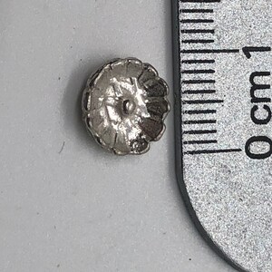 May include: A silver-toned metal button with a decorative, flower-like design. The button is approximately 1 cm in diameter.