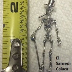 May include: Silver skeleton charm with a top hat and a pipe, holding a cane. The charm is labeled "El Samedi Calaca".