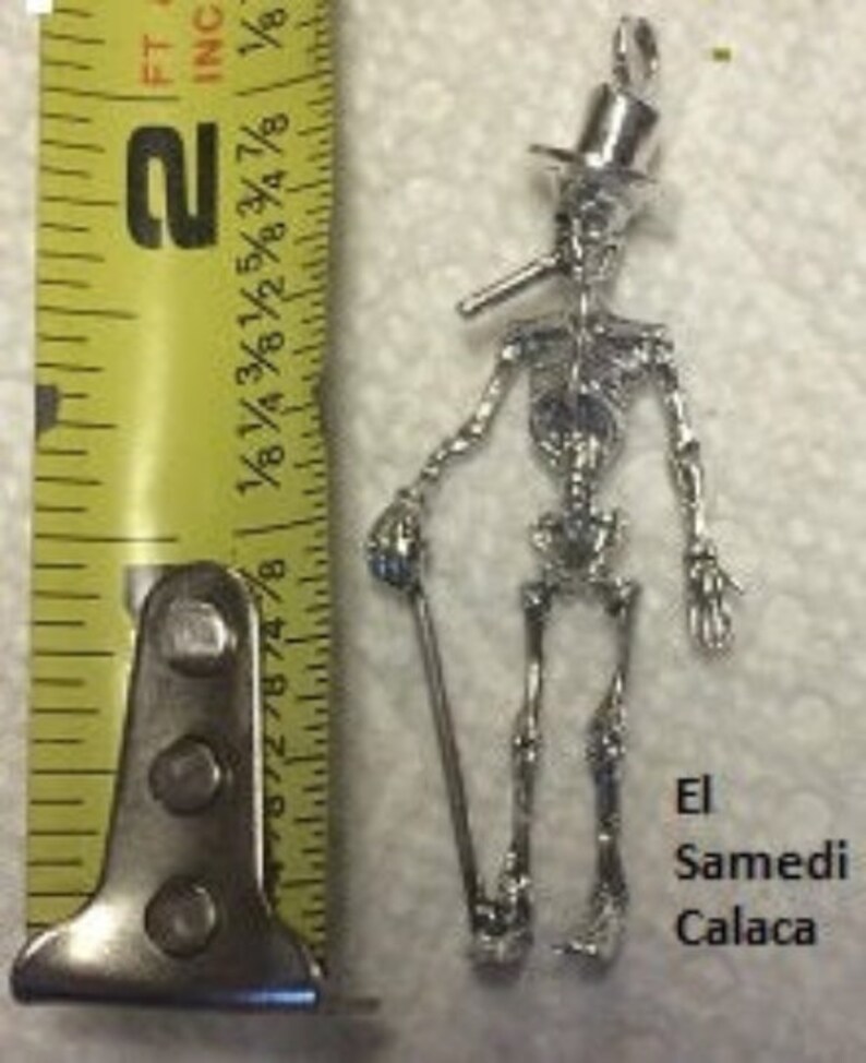 May include: Silver skeleton charm with a top hat and a pipe, holding a cane. The charm is labeled "El Samedi Calaca".
