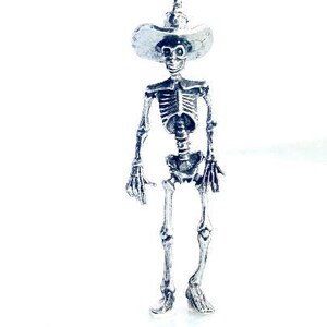 May include: A silver skeleton figurine wearing a wide-brimmed hat. The skeleton is standing with its arms at its sides and its legs slightly apart.