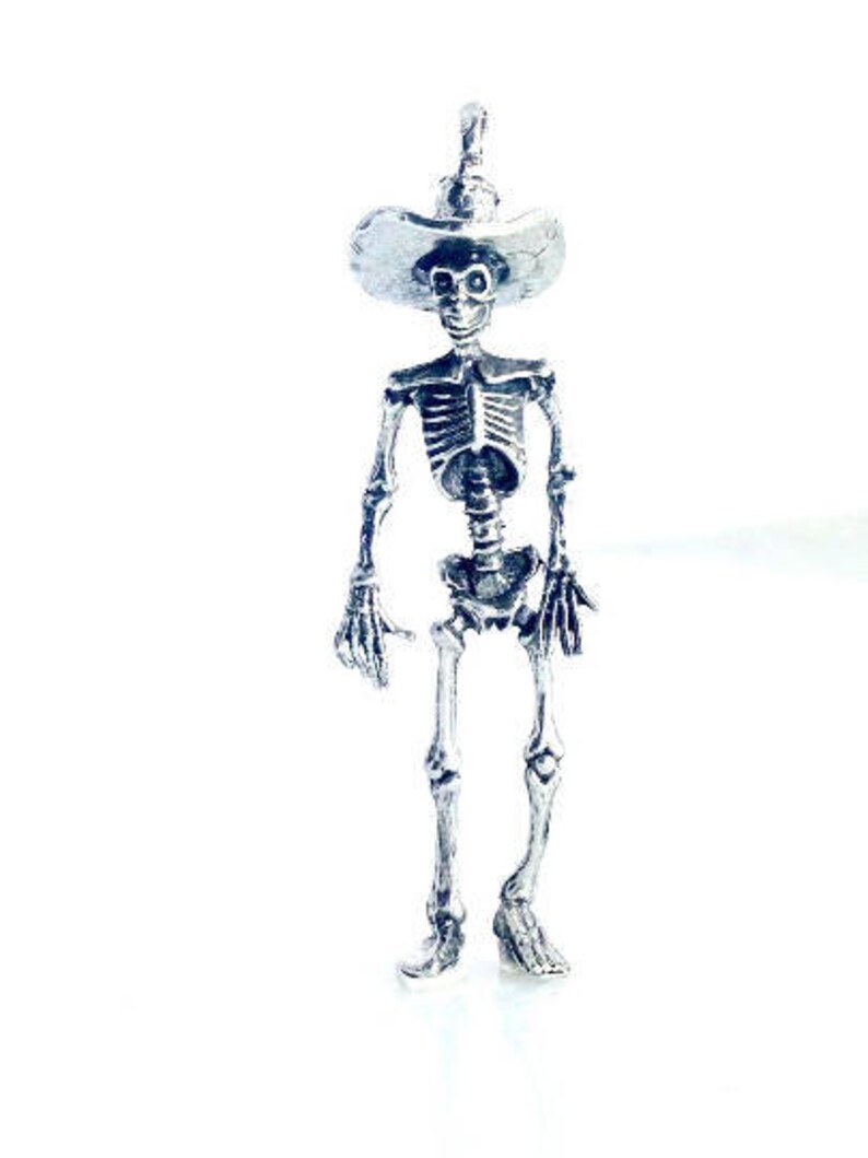 May include: A silver skeleton figurine wearing a wide-brimmed hat. The skeleton is standing with its arms at its sides and its legs slightly apart.