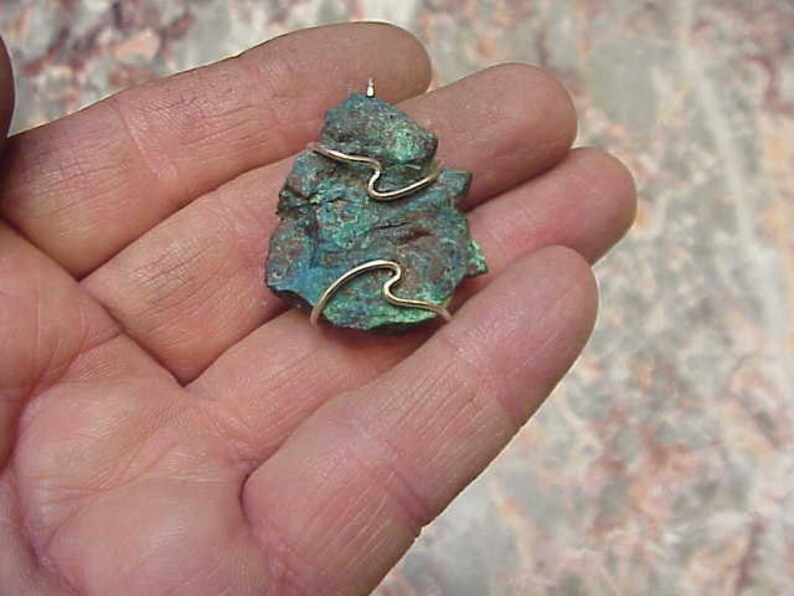 Puede incluir: Un colgante de piedra verde y marr&oacute;n con un envoltorio de alambre dorado. La piedra tiene una forma irregular y una textura rugosa.