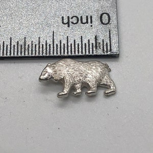 May include: A silver charm in the shape of a bear. The bear is walking and has a textured surface.