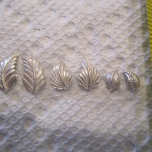 May include: Seven silver leaf charms of varying sizes, arranged in a row on a white surface.