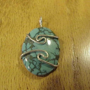 Puede incluir: Un colgante de piedra turquesa con envoltura de alambre plateado. La piedra preciosa tiene un patr&oacute;n de m&aacute;rmol y es de forma ovalada.