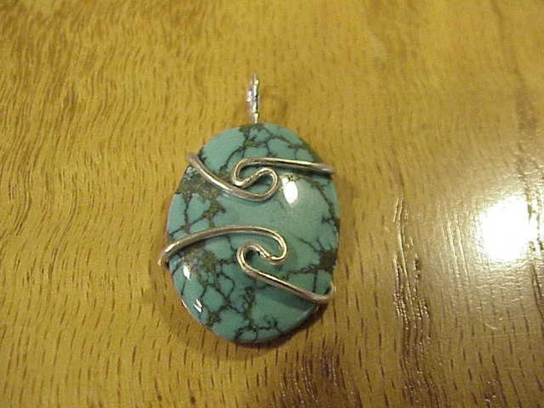 Puede incluir: Un colgante de piedra turquesa con envoltura de alambre plateado. La piedra preciosa tiene un patr&oacute;n de m&aacute;rmol y es de forma ovalada.