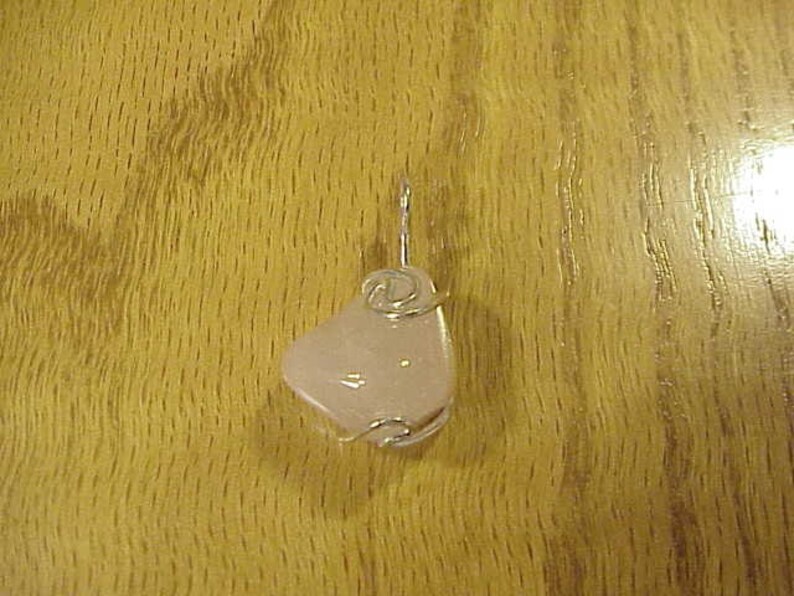 Puede incluir: Un colgante de coraz&oacute;n rosa con un envoltorio de alambre plateado. El colgante est&aacute; sobre una superficie de madera.