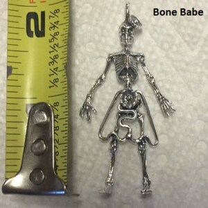 May include: Silver skeleton charm with a detailed anatomical design, including internal organs. The charm is approximately 2 inches tall and features the text "Bone Babe".
