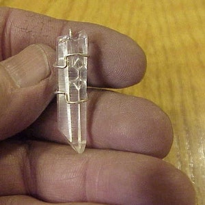Puede incluir: Un colgante de cristal de cuarzo transparente con un envoltorio de alambre dorado. El cristal es puntiagudo en la parte inferior y tiene una textura natural y rugosa.