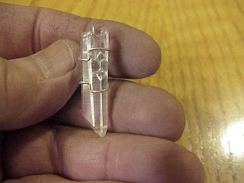Puede incluir: Un colgante de cristal de cuarzo transparente con un envoltorio de alambre dorado. El cristal es puntiagudo en la parte inferior y tiene una textura natural y rugosa.