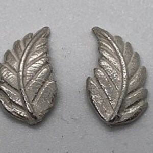 May include: Two silver-colored leaf-shaped earrings. Each earring has a detailed, textured surface with visible veins, resembling a feather. The earrings are displayed on a light gray surface, showcasing their intricate design and metallic sheen.