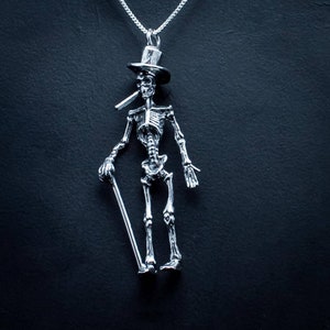 May include: A silver skeleton pendant necklace with a top hat and a cane. The skeleton is wearing a top hat and holding a cane. The necklace is on a black background.