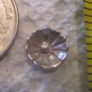 May include: A silver-colored metal flower-shaped button with a raised center. The button has a scalloped edge and a slightly textured surface.