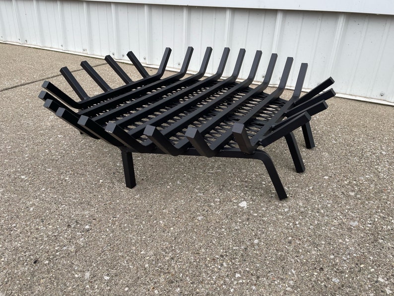Op de afbeelding: Zwarte metalen haardrooster met een roosterontwerp en afgeschuinde poten. Het rooster is ontworpen om houtblokken voor een vuur te houden.