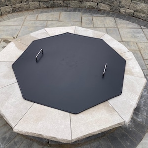 May include: Octagonal black metal fire pit cover with two silver handles. The cover is sitting on a stone fire pit.