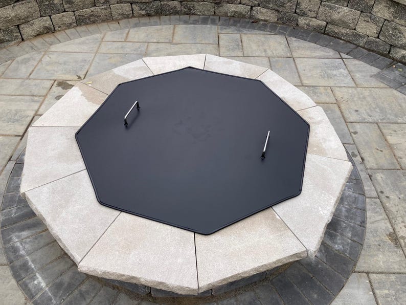 May include: Octagonal black metal fire pit cover with two silver handles. The cover is sitting on a stone fire pit.