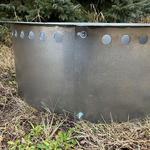 May include: A circular galvanized steel fire pit ring with a metallic silver finish. The structure features a series of evenly spaced circular ventilation holes along the upper edge and is secured with visible metal bolts at the vertical seam.