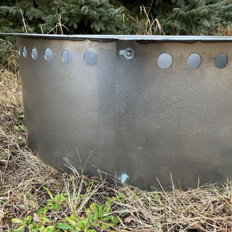 May include: A circular galvanized steel fire pit ring with a metallic silver finish. The structure features a series of evenly spaced circular ventilation holes along the upper edge and is secured with visible metal bolts at the vertical seam.