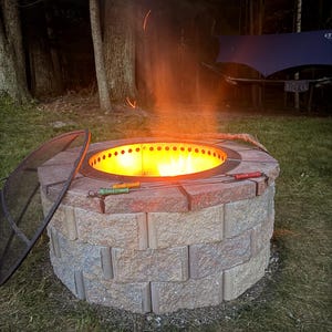 May include: A circular fire pit constructed from stacked, textured tan and reddish-brown concrete blocks. A metal insert with ventilation holes contains a bright orange flame. A black mesh spark screen rests nearby, and two metal roasting skewers lie across the top.
