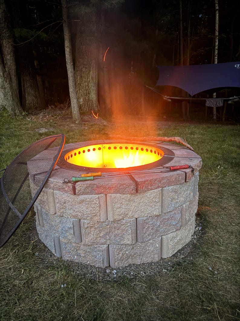 May include: A circular fire pit constructed from stacked, textured tan and reddish-brown concrete blocks. A metal insert with ventilation holes contains a bright orange flame. A black mesh spark screen rests nearby, and two metal roasting skewers lie across the top.
