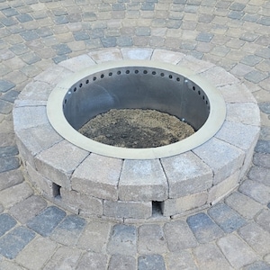 May include: A circular fire pit constructed from gray concrete pavers arranged in a radial pattern. The center features a metallic ring insert with a series of ventilation holes along the top edge, surrounding a base filled with dark soil and ash.