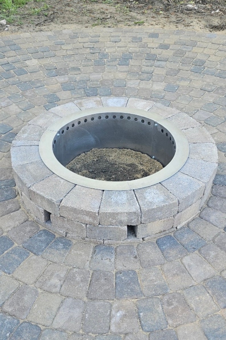 May include: A circular fire pit constructed from gray concrete pavers arranged in a radial pattern. The center features a metallic ring insert with a series of ventilation holes along the top edge, surrounding a base filled with dark soil and ash.