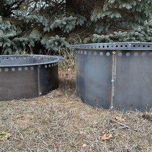 May include: Two circular fire pit rings constructed from dark grey steel. Each ring features a flat, outward-extending rim at the top with a series of evenly spaced circular ventilation holes. The metal panels are joined by visible vertical bolts.