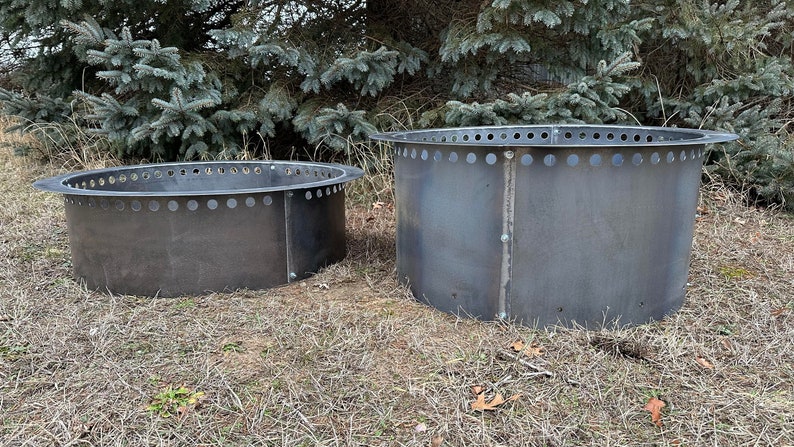 May include: Two circular fire pit rings constructed from dark grey steel. Each ring features a flat, outward-extending rim at the top with a series of evenly spaced circular ventilation holes. The metal panels are joined by visible vertical bolts.