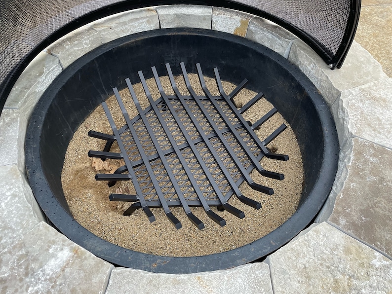 Op de afbeelding: Een zwarte metalen vuurkorf rooster met een zeshoekige vorm en een gaasontwerp. Het rooster zit in een ronde vuurkorf gevuld met zand.