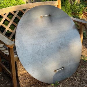 May include: A large, round, gray metal lid with two silver handles. The lid is resting on a wooden bench with a lattice back. The bench is outdoors, surrounded by greenery.