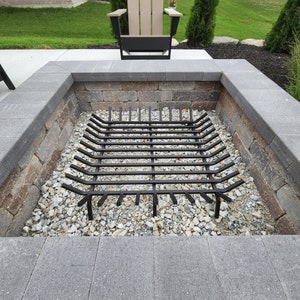 May include: A black metal fire pit grate sits inside a stone fire pit. The fire pit is surrounded by gray pavers and a small bed of gravel.